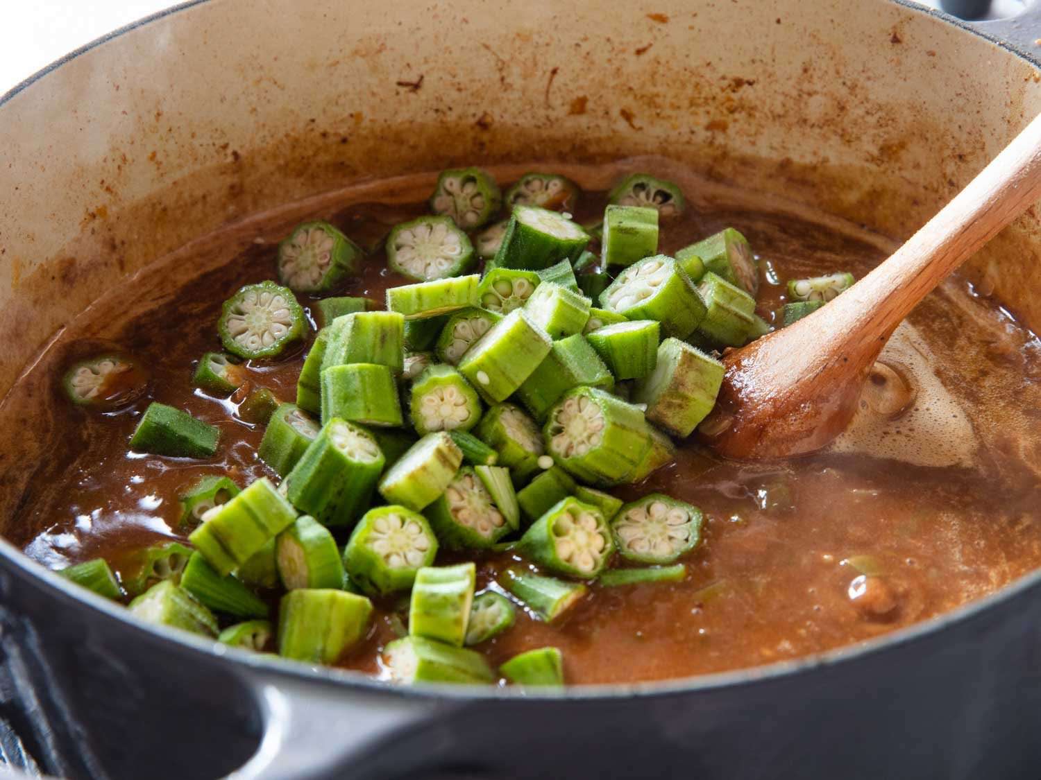 Adding okra to the pot of gumbo and stirred in with a wooden spoon.