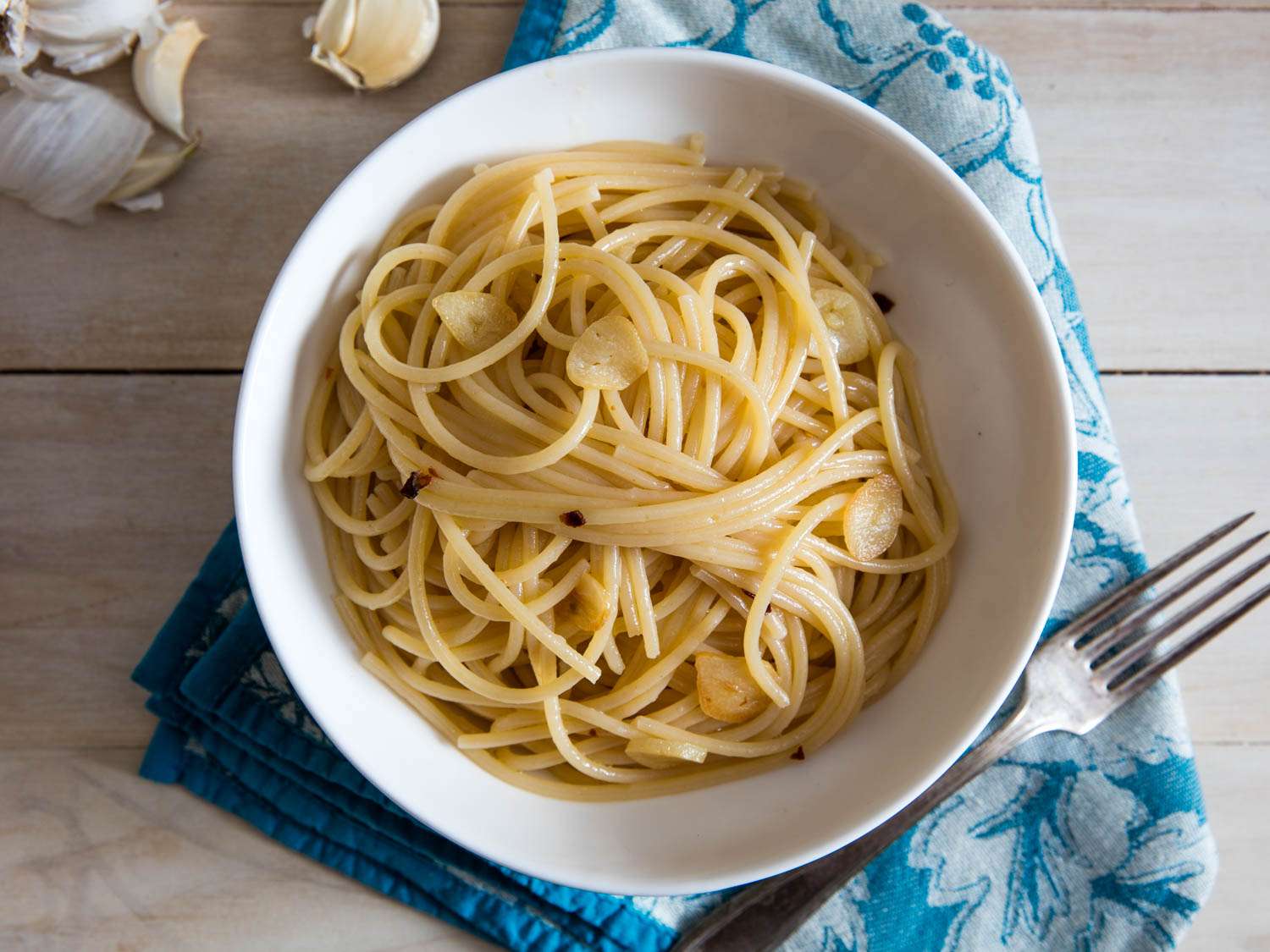 An overhead view of pasta aglio e olio in a white bowl next to a fork, garlic cloves, and a blue and white floral napkin.