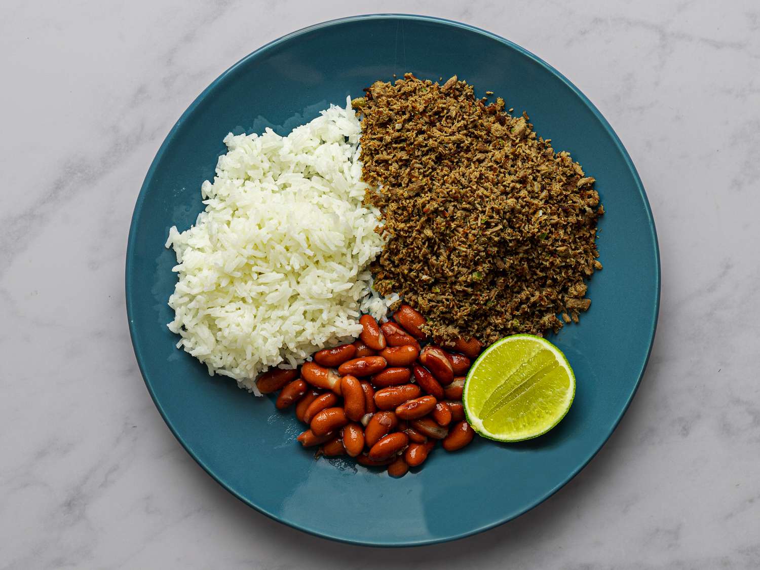 SalpicÃ³n, white rice, red kidney beans, and a slice of lime on a blue plate on a marble counter.
