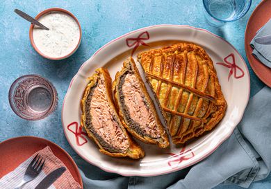 Salmon encased in pastry on a serving dish with dill sauce and tableware on a blue surface