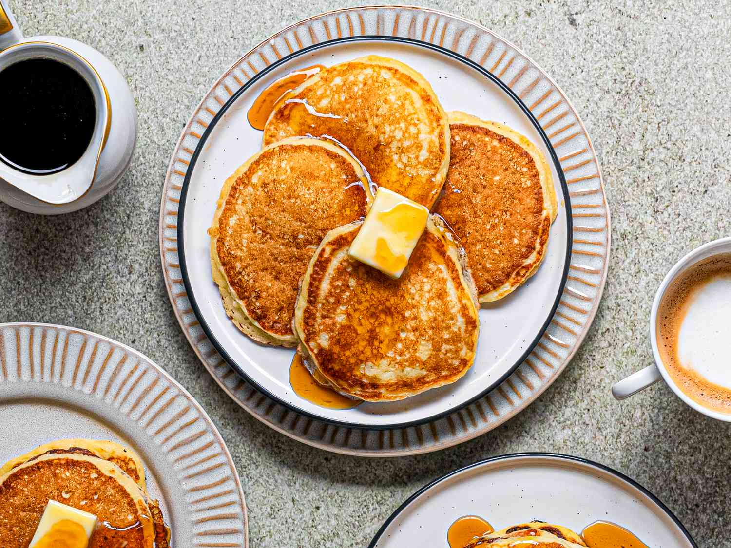 Plate of extra light and fluffy buttermilk pancakes topped with pat of butter and drizzled with maple syrup displayed against a grey speckled background