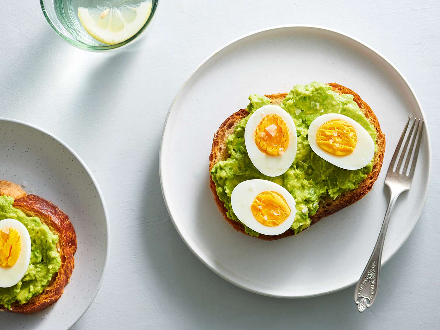 Halved hard-boiled eggs topped with flaky salt and placed on bread topped with mashed avocado. The food is placed on a white ceramic plate with a metal fork.