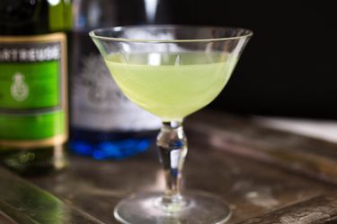 A Last Word cocktail in a stemmed coupe glass with an out-of-focus bottle of green Chartreuse and gin in the background.