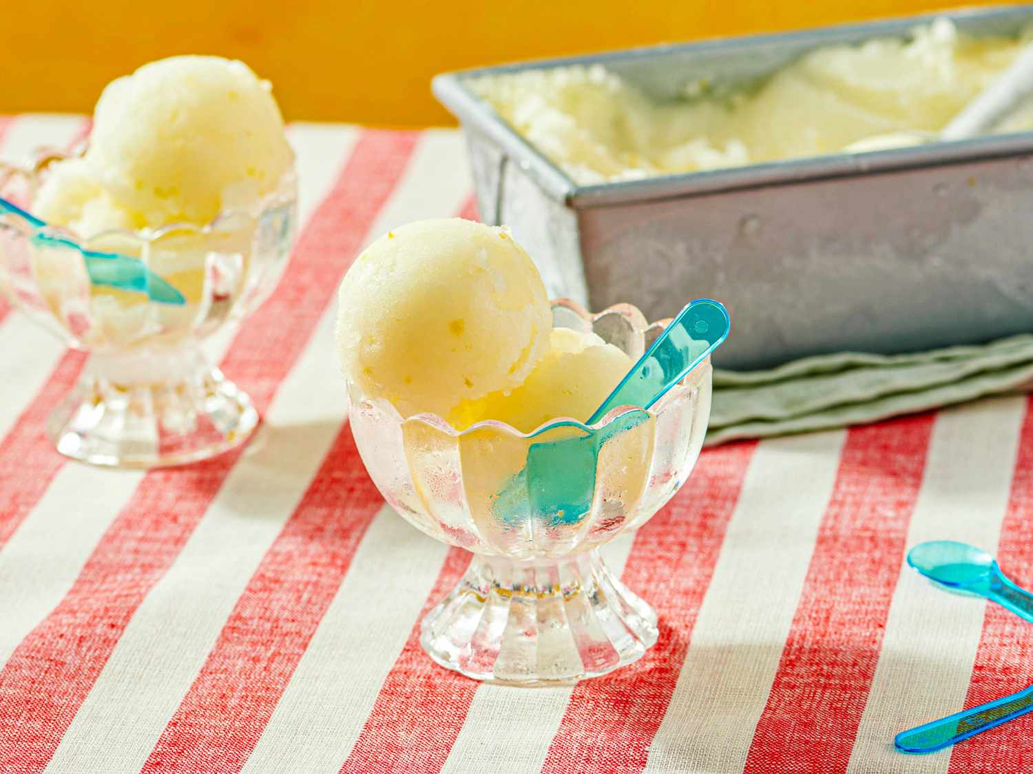 Two dessert bowls with scoops of lemon sorbet each with a small plastic spoon on a striped tablecloth with a tray in the background