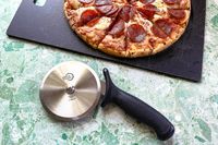 A pepperoni pizza partially sliced on a cutting board lying next to a Mercer pizza cutter with a textured handle on a green marble countertop