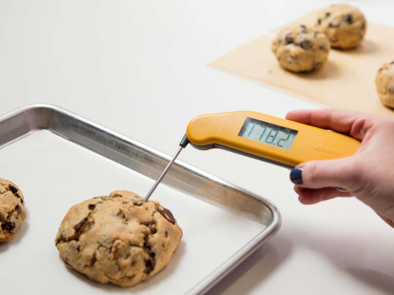 Using a probe thermometer to take the temperature of a chocolate chip cookie on a parchment-lined baking sheet. 