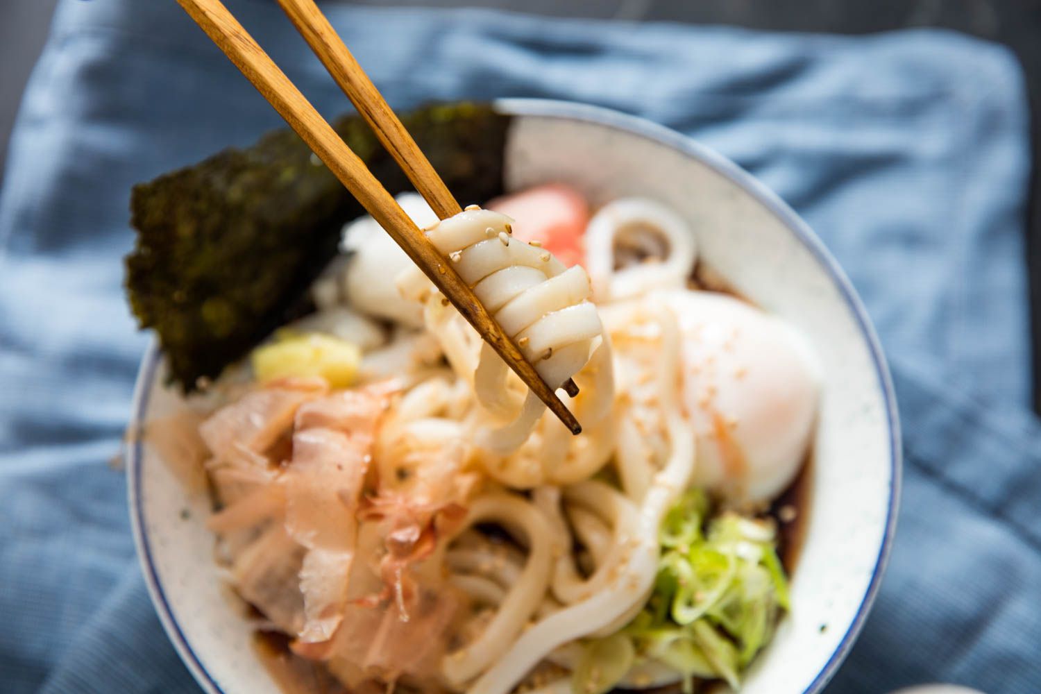 A close-up of several udon noodles being lifted above a bowl of bukkake udon with a pair of chopsticks.