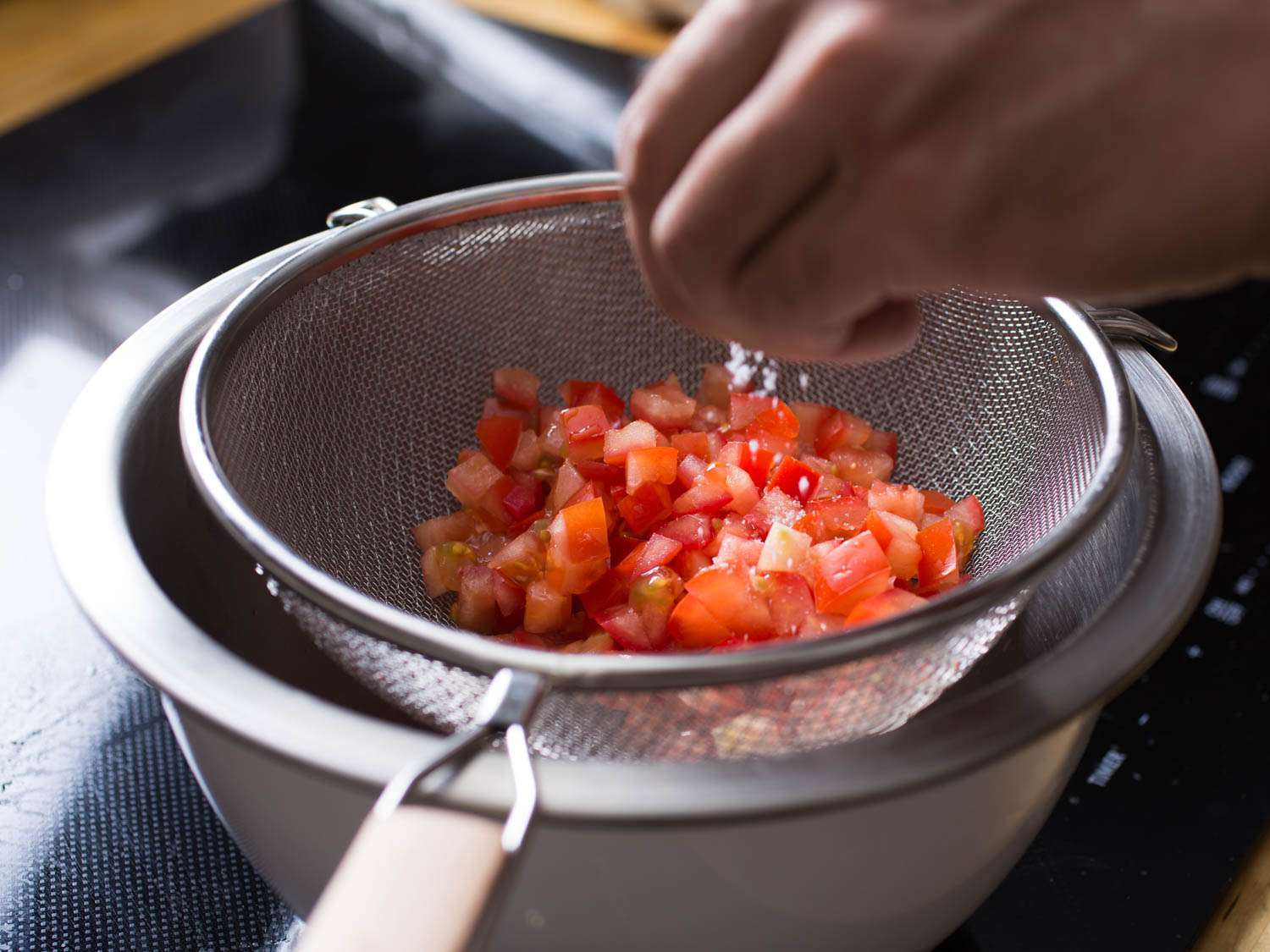 Diced tomatoes on a strainer being sprinkled with salt.
