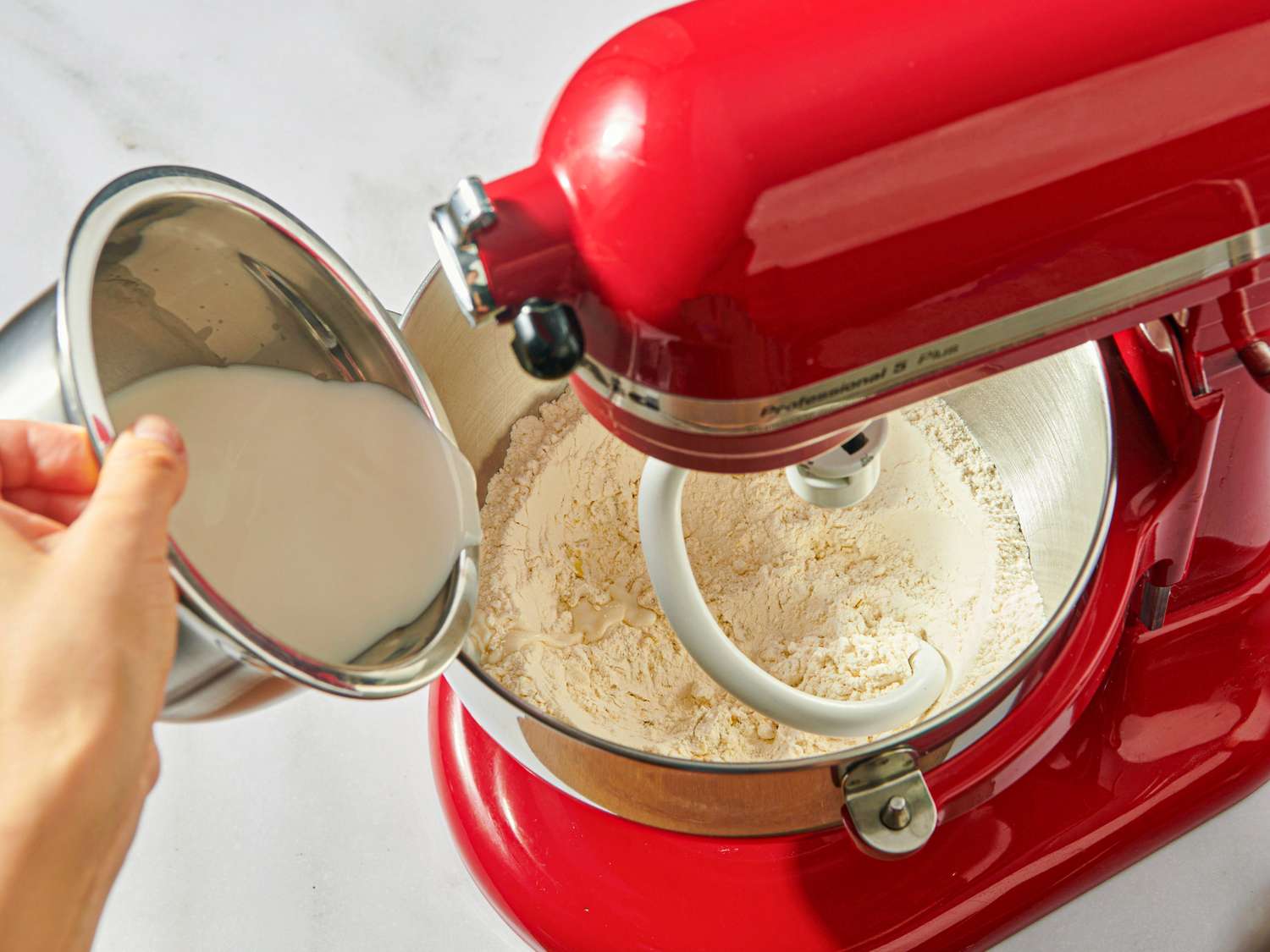 Person pouring an ingredient into a red stand mixer with a dough hook attachment