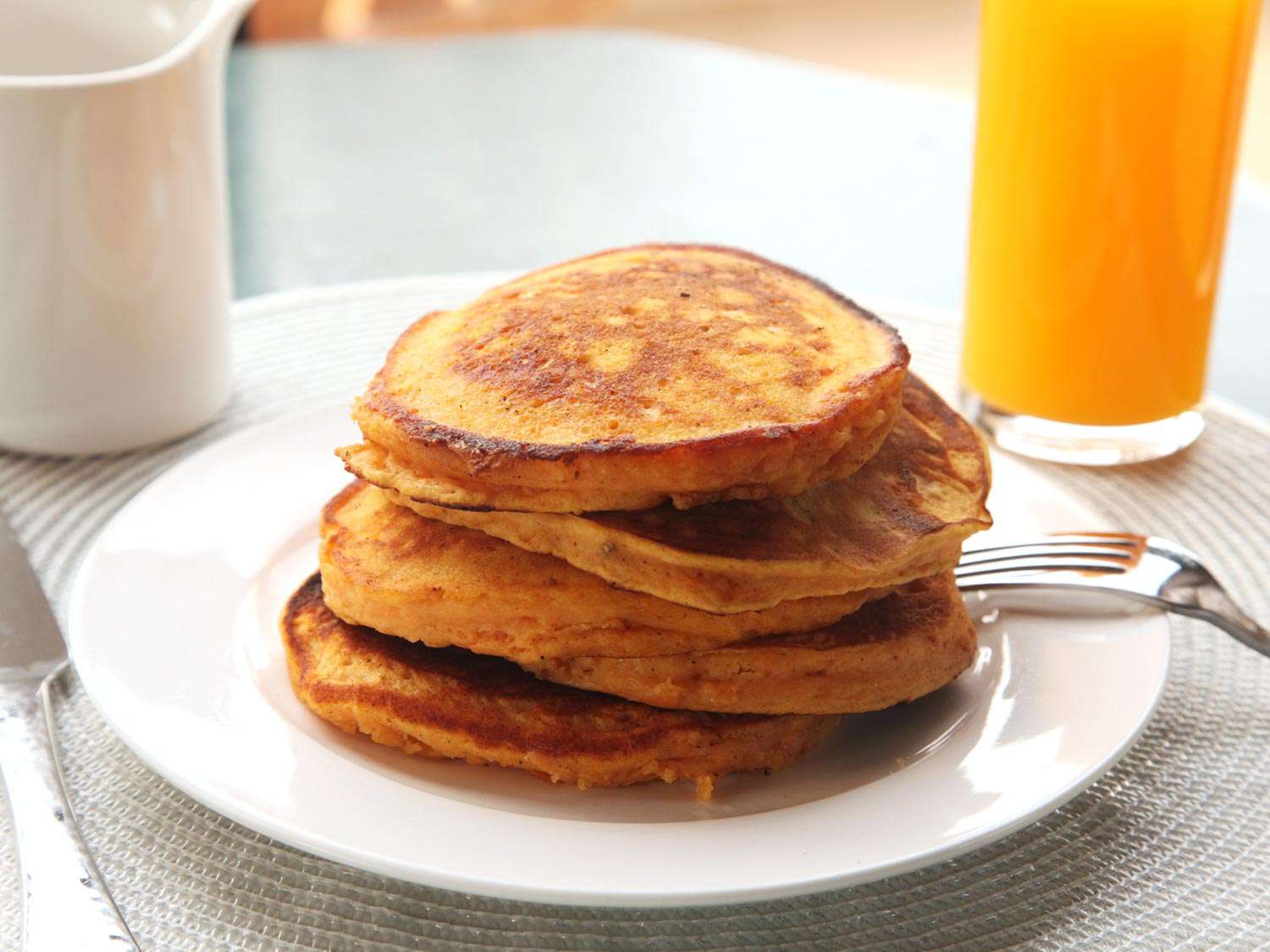 A stack of sweet potato pancakes on a white plate with a fork next to a glass of orange juice and a pitcher of maple syrup.