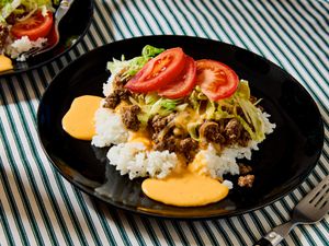 A plate of taco rice with beef, lettuce, and sliced tomatoes on top of white rice, served on a striped tablecloth