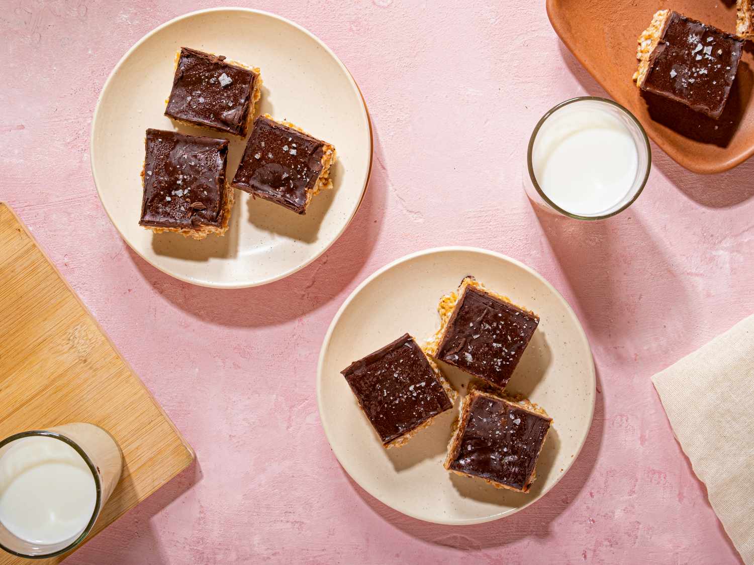 Plates of scotcheroos, chocolate-topped dessert bars, placed on a pink surface beside glasses of milk