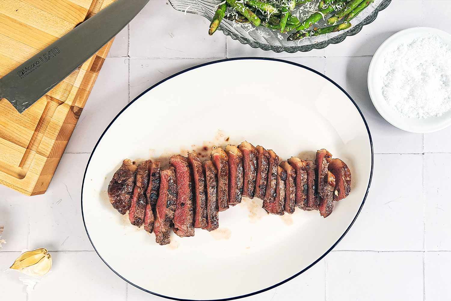 Sliced cooked steak on a white plate kitchen tools and side dishes visible nearby