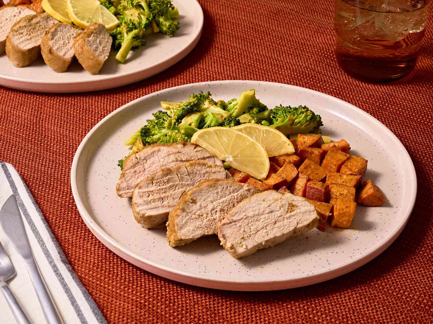 Slices of pork, vegetables and lemon slices on white plates, with a white napkin, and a fork and knife on the side. Settings sot o top of red tablecloth. 