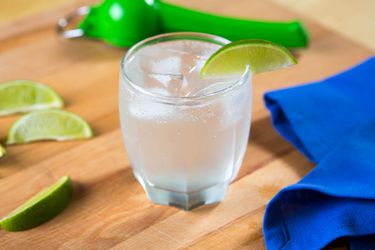 A gin rickey in a highball glass with a lime wedge on the rim. The glass is on a wood cutting board with a blue napkin, three other lime wedges, and a green hinged citrus juicer. 