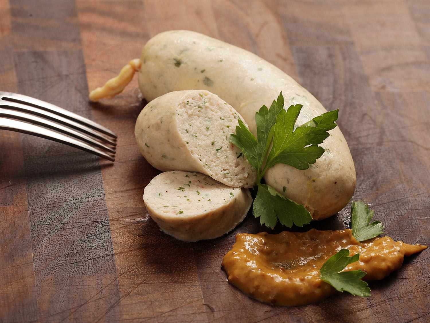 Closeup of weisswurst arranged on a cutting board with a smear of mustard and a few parsley leaves.