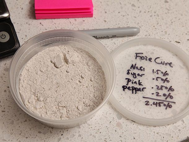 A half-pint deli container filled with the ground curing mixture. The lid has the ingredients and their percentages by weight written in permanent marker.