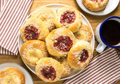 Kolaches on a platter, with one cream and one strawberry on 2 plates, with coffee on the side, striped napkins, on a wooden surface. 
