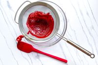 A red spatula beside the Rösle Stainless Steel Round Handle Kitchen Strainer with red puree on a clean surface