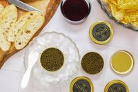 A spread featuring a bowl of caviar on ice slices of bread a glass of red wine chips and goldlabeled caviar tins