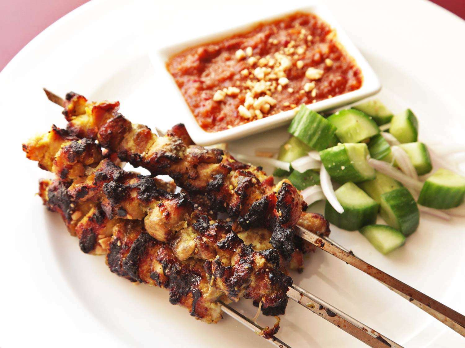Three skewers of Thai-style chicken satay on a serving plate with cucumbers and peanut tamarind dipping sauce. 