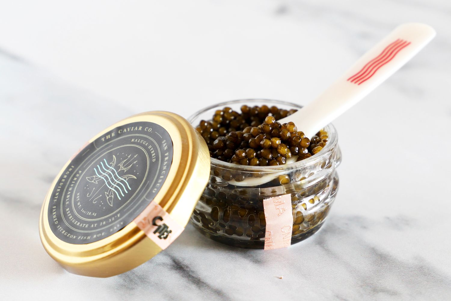 An open jar of Caviar Co kaluga hybrid caviar with a mother-of-pearl spoon inside