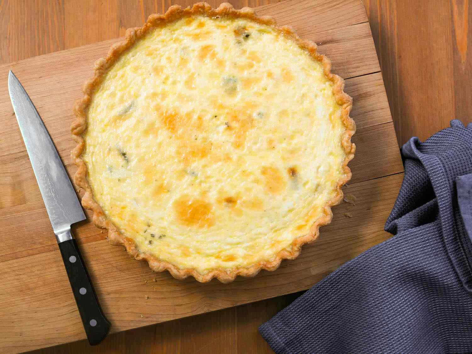A cheese quiche on a wooden cutting board with a knife and a kitchen towel. 