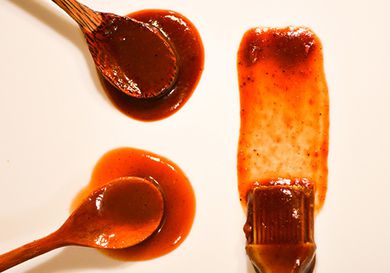Overhead image of two wooden spoons and a brush that have been doused in barbecue sauce and arranged on an off-white surface.