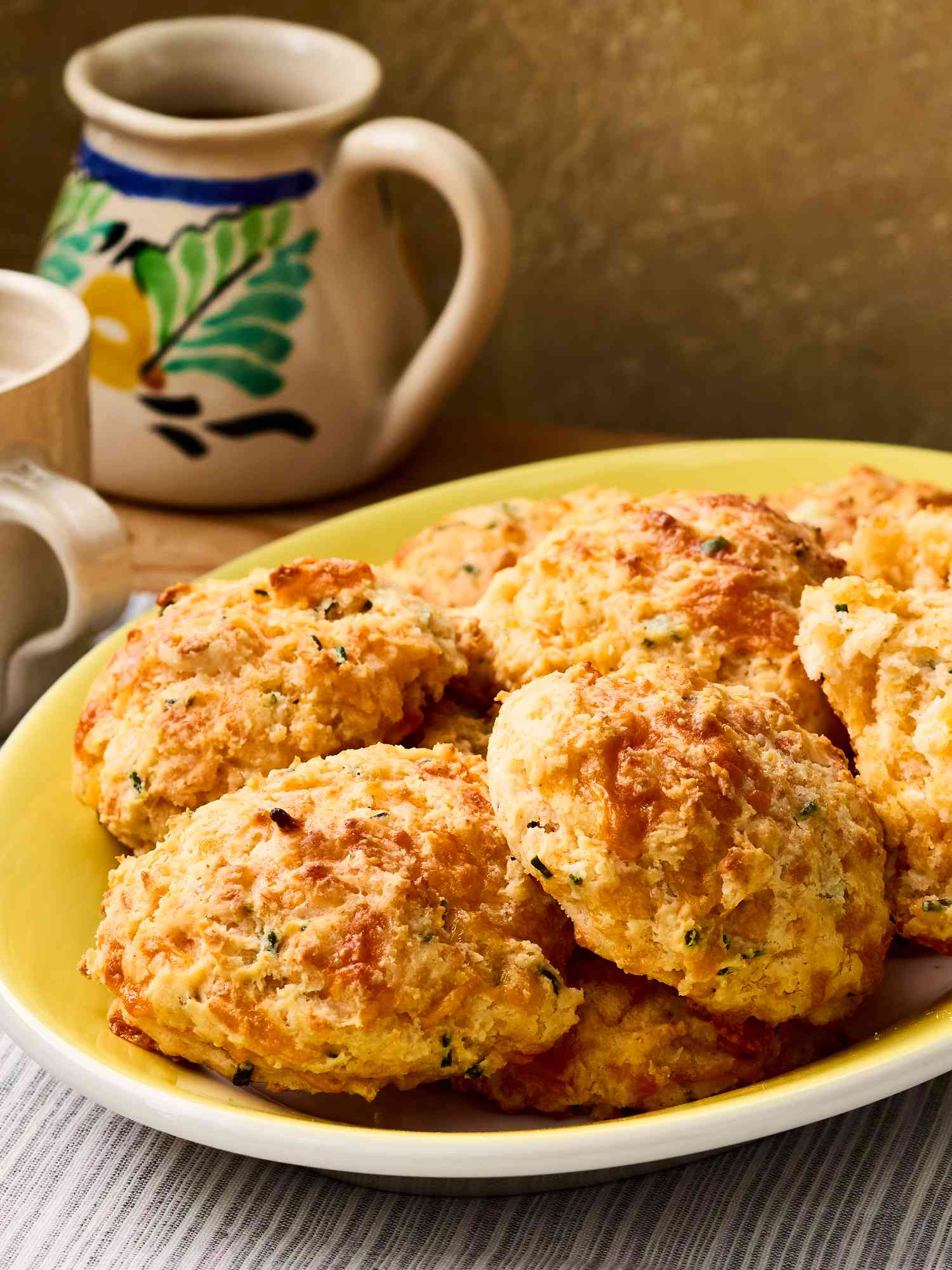 A plate of cheddar chive biscuits, placed on a table with a decorative pitcher in the background