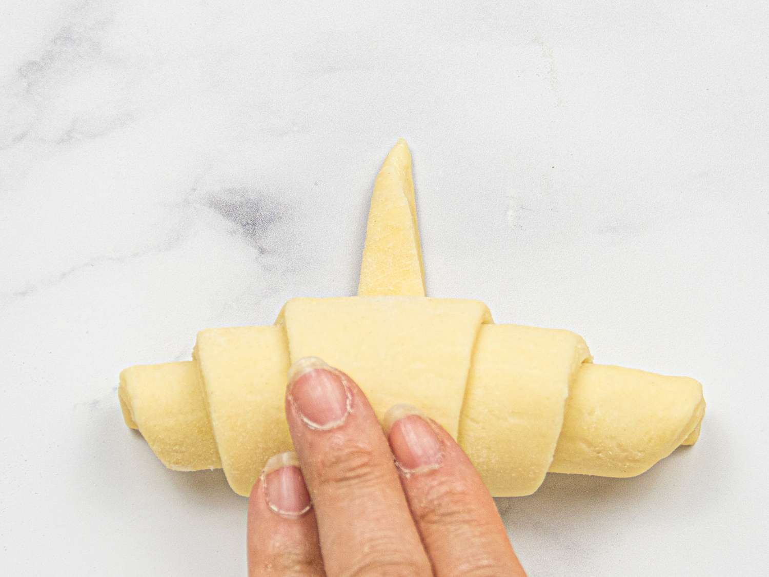 A persons hand shaping a croissant dough on a white surface