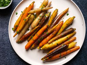 A round white porcelain plate holding multi-colored sous vide glazed carrots. The plate is on a dark blue textured surface and there is a small white bowl holding chopped parsley in the top left corner of the image.