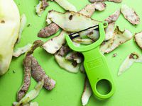 A green y-peeler on a green cutting board surrounded by potato skins and a peeled potato to the side of it