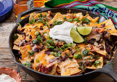 A skillet of nachos topped with melted cheese, black beans, sour cream, lime wedges, and green onions, placed on a wooden surface