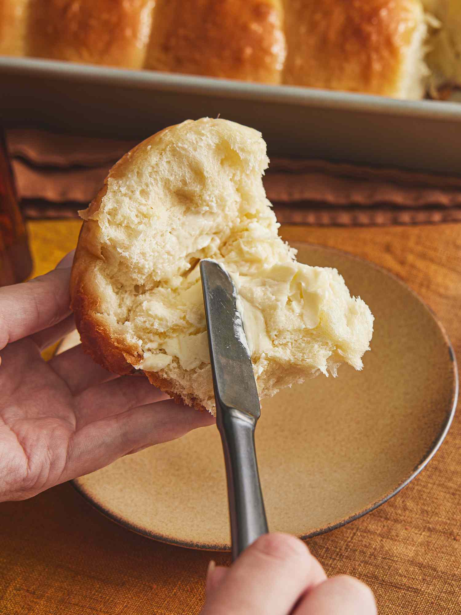 Parkerhouse roll ripped open with butter being spread on it. The table has a small plate on it, a plate of butter, and linen table cloth.