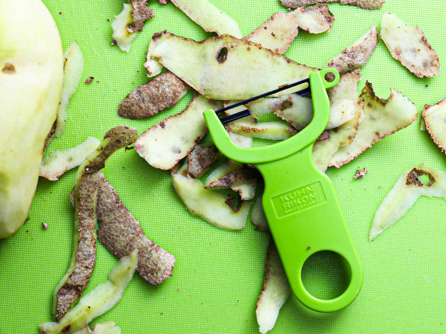 A green y-peeler on a green cutting board surrounded by potato skins and a peeled potato to the side of it