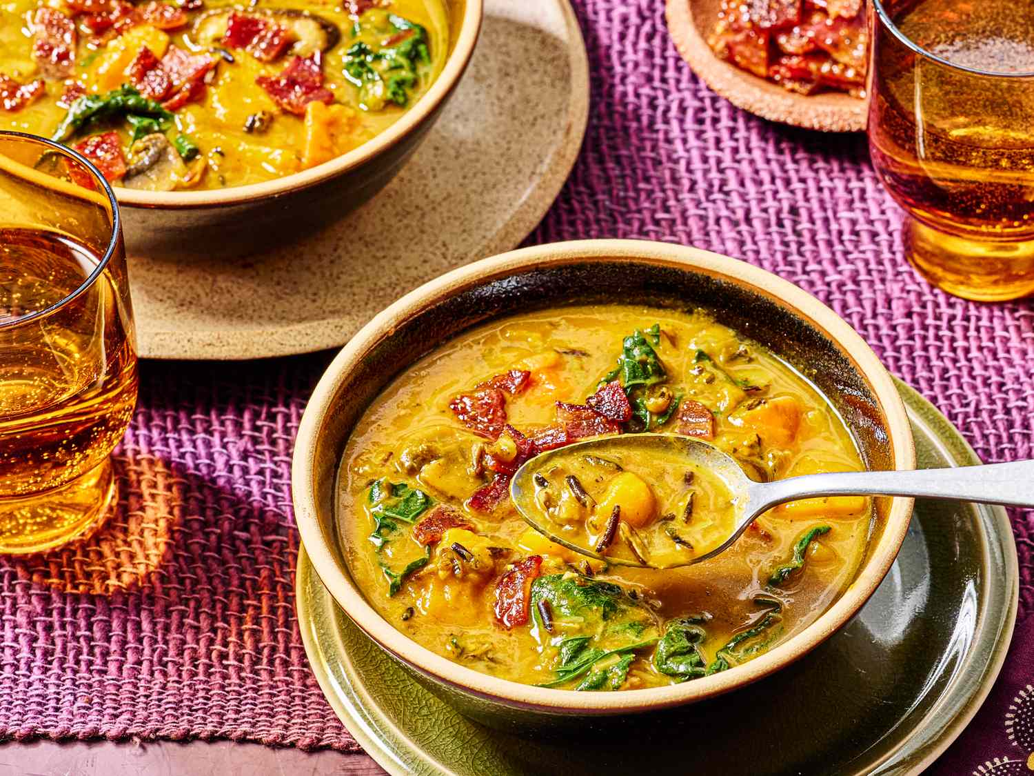 Bowls of pumpkin soup with wild rice and greens spoons placed in one bowl glasses of drink nearby