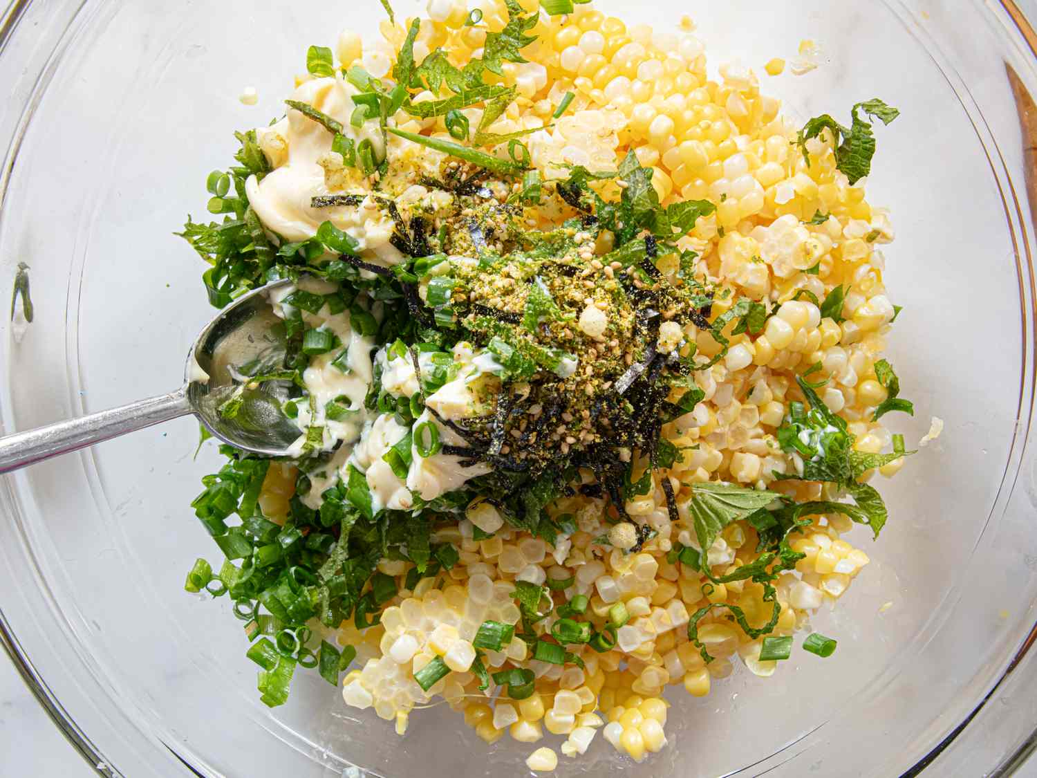 A bowl containing corn kernels mixed with herbs and seasoning with a spoon visible in the mixture