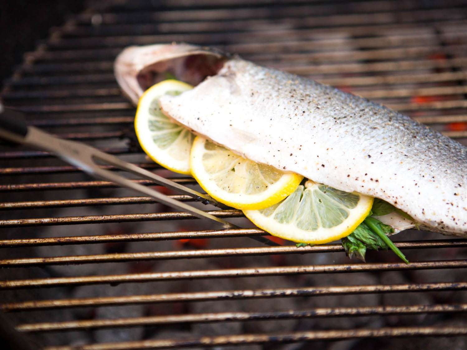 Author positions the tines of a carving fork between the grates and underneath a fish to flip it on the grill.