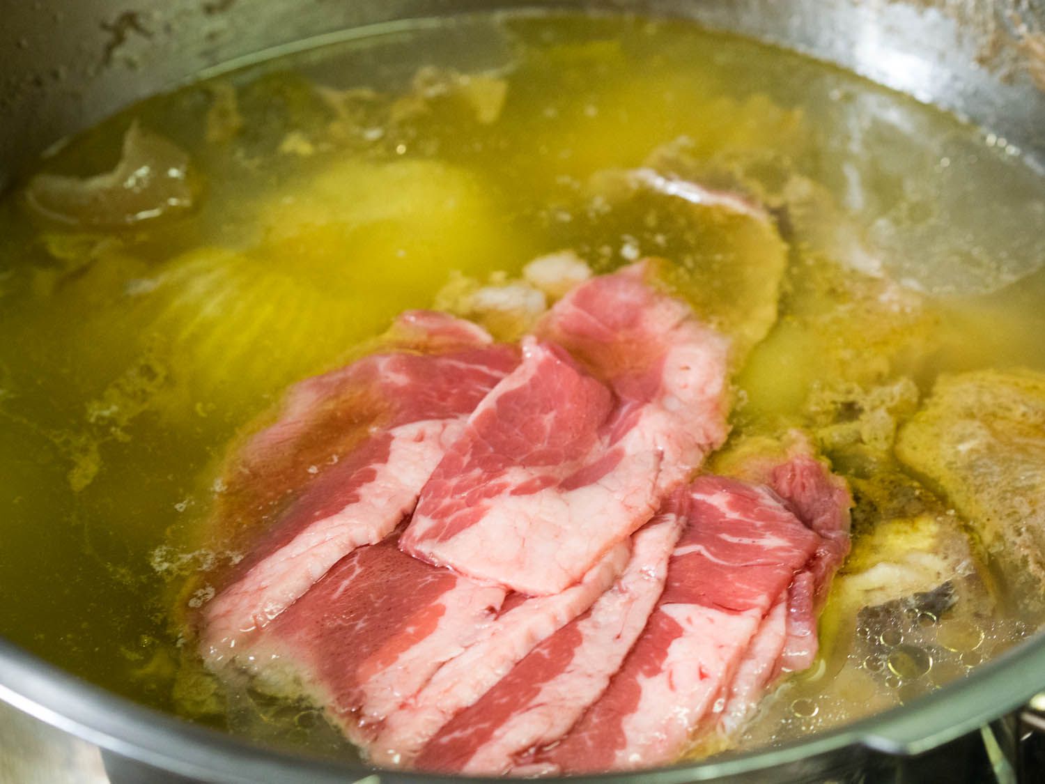 A stainless steel pot holding chicken broth cooked with vegetables, with some thinly sliced, uncooked beef on top.