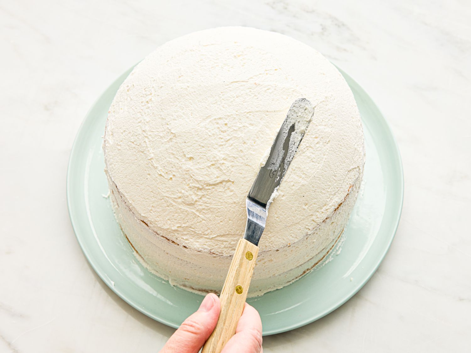 A hand using a spatula to frost a round cake on a plate