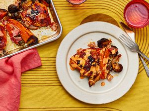 Serving of sheet-pan chicken and vegetables on a plate with a fork and knife and a red cup. To the side of the dish is the tray of chicken and vegetables with a red napkin
