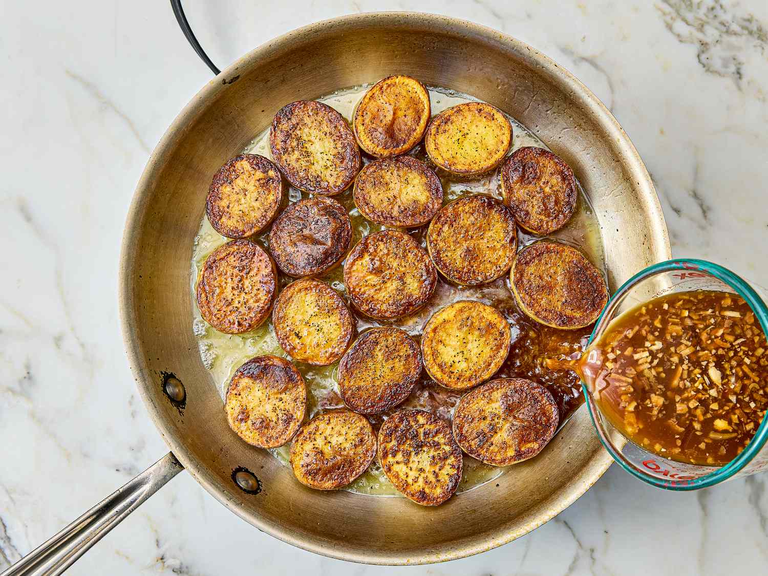 Panfried potato slices with a bowl of onion broth being poured in in a skillet on a marble surface