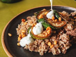 A dish with stuffed zucchini rice and yogurt sauce being poured on top garnished with pine nuts
