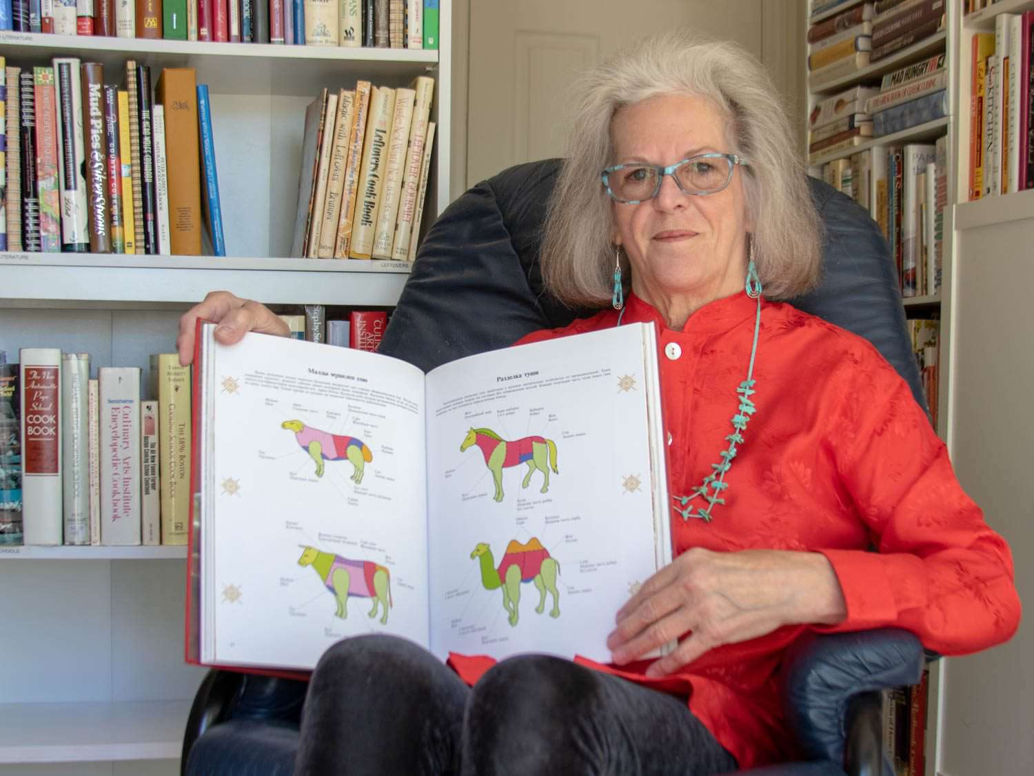 Cookbook collector Sue Jimenez, seated, holding open a cookbook to the camera, with books on shelves behind her.