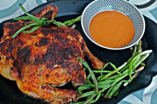 A whole Spanish roast chicken on a platter, garnished with whole scallions and a dish of romesco sauce