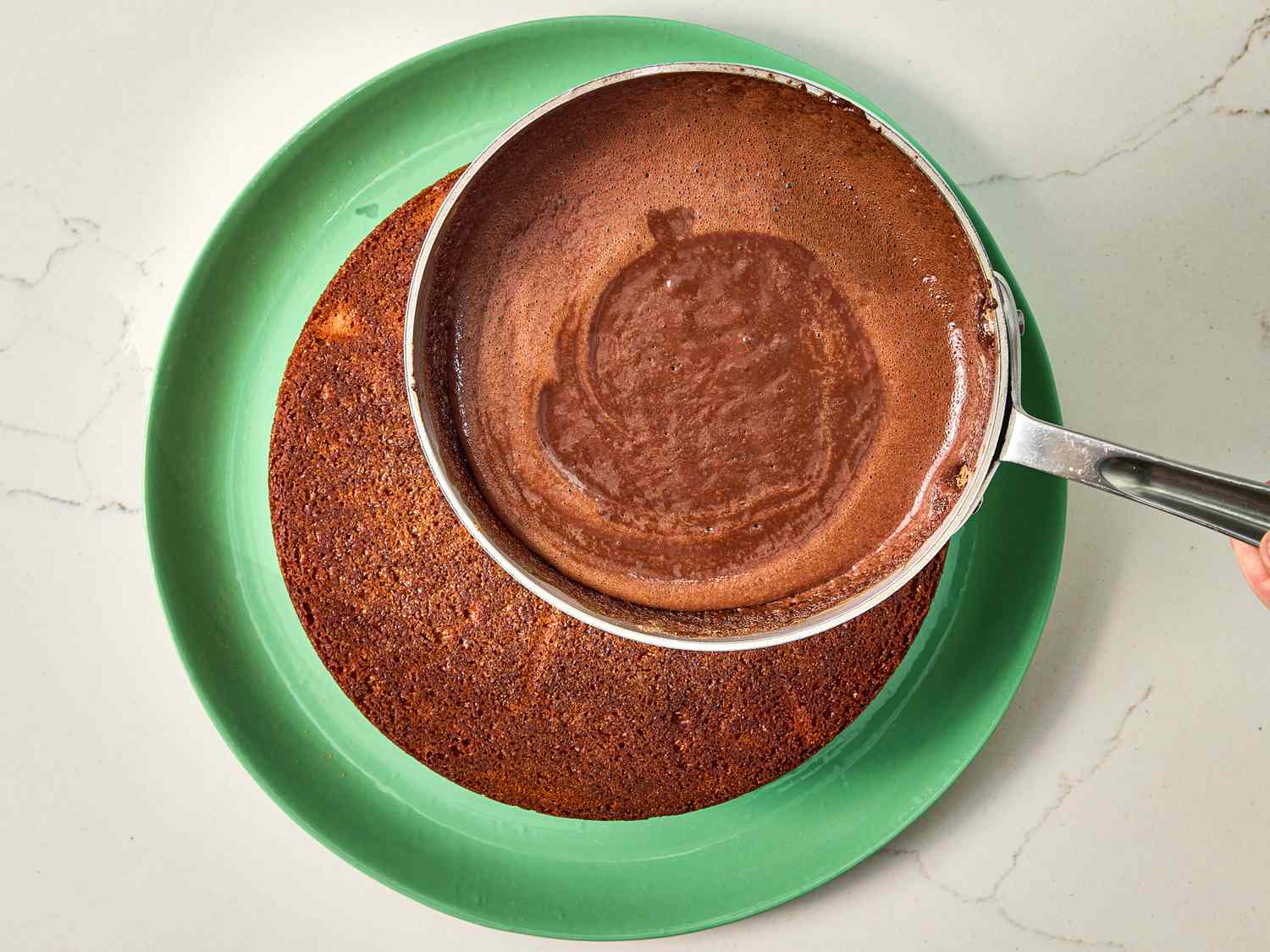 A chocolate glaze being poured from a saucepan onto a round cake placed on a green plate