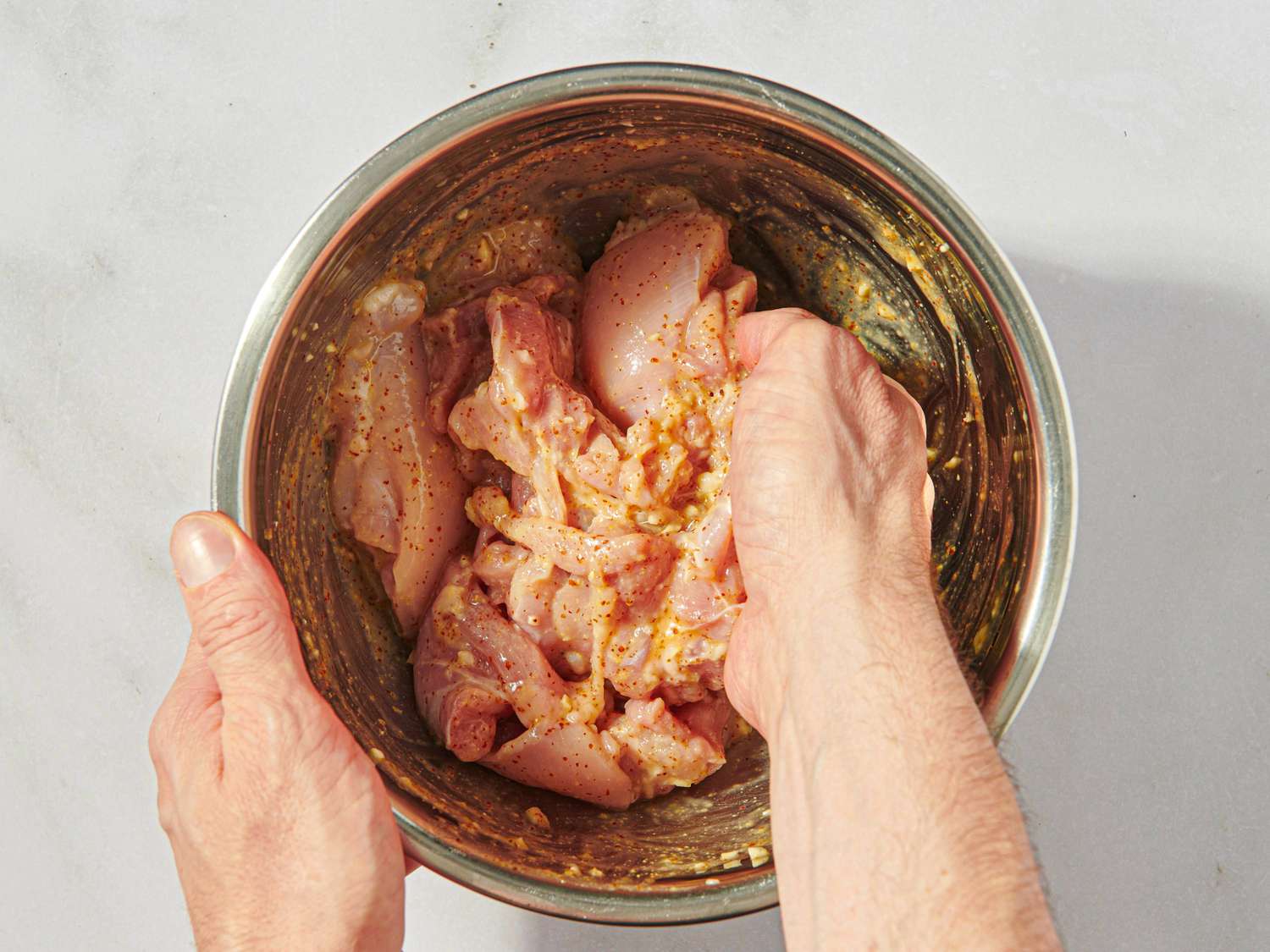 Hands mixing chicken pieces with marinade in a metal bowl