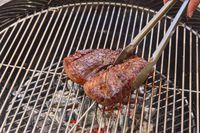 A hand using tongs to move steak on the Weber Original Kettle Premium 22-Inch Charcoal Grill