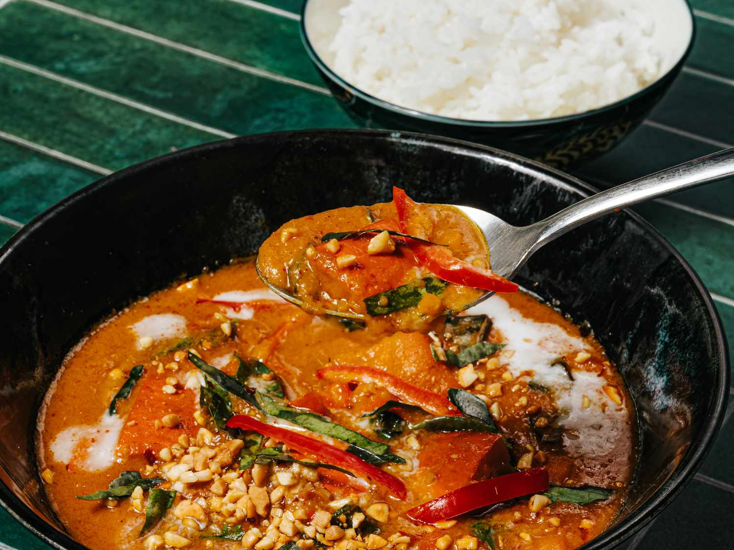 Fully composed Squash curry with basil and chili toppings, in a black bowl, on a deep green tile surface. In the top corner is a bowl of white rice