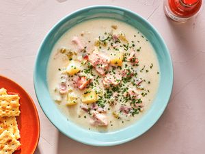 A baby blue bowl holding a serving of creamy salmon chowder. The chowder is topped with minced herbs and there is a red plate of saltine crackers in the bottom left corner of the image, and a bottle of Tabasco hot sauce in the top right corner of the image.